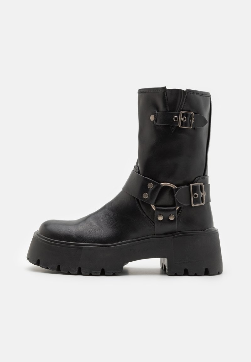 Botín de cuero negro hasta el tobillo con una suela gruesa, que presenta dos correas similares a cinturones con hebillas plateadas y un acento de anillo circular.