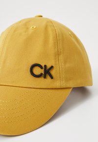 Gul bomullskeps med en böjd skärm, prydd med en svart broderad "CK"-logotyp framtill. Textil med detaljerad sömnad.