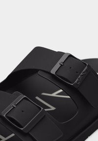 Μαύρο σανδάλι με δύο ρυθμιζόμενους ιμάντες με αγκράφες και μερικώς ορατό λευκό λογότυπο της μάρκας στο πέλμα, απομονωμένο σε φωτεινό φόντο.