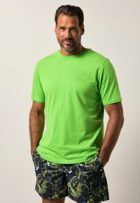 T-shirt verde brillante con maniche corte e collo rotondo, caratterizzato da un logo circolare testurizzato. Abbinato a pantaloni corti scuri con motivo floreale.
