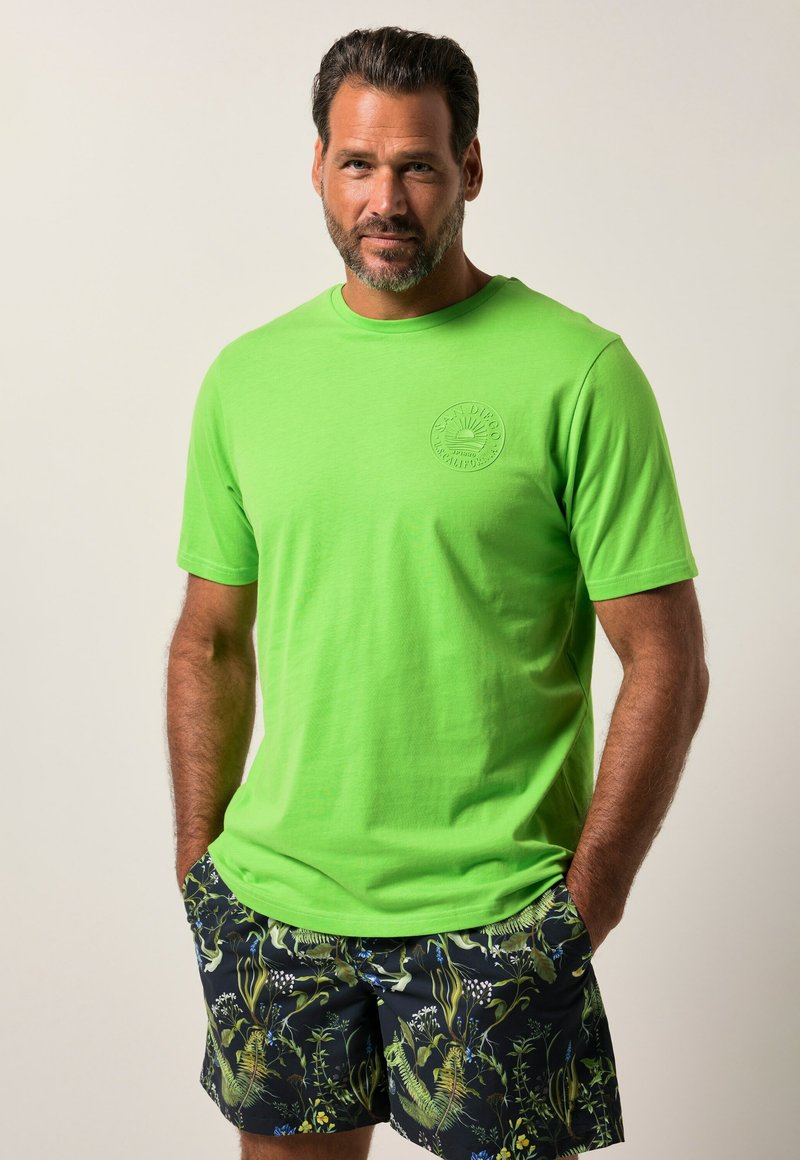 T-shirt verde brillante con maniche corte e collo rotondo, caratterizzato da un logo circolare testurizzato. Abbinato a pantaloni corti scuri con motivo floreale.