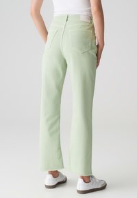 Jeans verde chiaro a gamba dritta, con orlo sfrangiato e due tasche posteriori, abbinate a sneakers bianche su uno sfondo neutro.