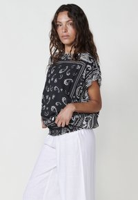 Blouse noire avec un motif paisley, manches courtes et ourlet élastique. Associée à un pantalon blanc ample en matériau léger.