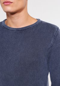 Nahaufnahme einer Person, die einen dreifarbig strukturierten Pullover in Marineblau mit einem gerippten Rundhalsausschnitt trägt.