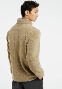 Beżowy polarowy sweter z wysokim kołnierzem, o fakturowanej powierzchni i luźnym kroju; posiada kontrastową niebieską podszewkę i boczne kieszenie.