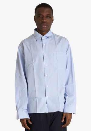 Giovane uomo che indossa una camicia oversize azzurro chiaro a righe sottili con tasca frontale e pantaloni scuri, in piedi su sfondo bianco.