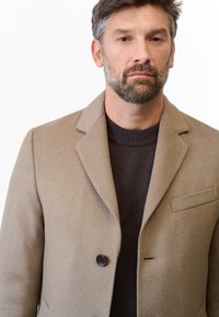 Beige ullkappa med notch-krage, en svart knapp och bröstficka. Buren över en mörk strukturerad tröja.