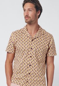 Camisa de manga corta con un patrón geométrico de zigzag en amarillo, marrón y crema. La tela es ligera, con un collar relajado y cierre de botones.