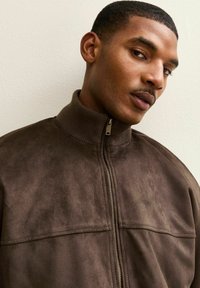 Bruin ritsjack gemaakt van een zacht suède-achtige stof, met een hoge kraag en twee borstzakken met een gladde textuur.