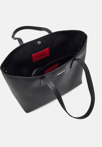 Musta nahkainen olkalaukku, jossa on teksturoitu pinta, leveät hihnat ja punainen sisämerkki. Varustettu napsautettavalla suljennalla ja runsaalla säilytystilalla.