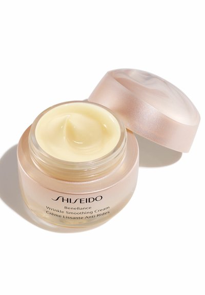 Tarro de vidrio con una tapa de color rosa, que contiene una crema alisadora de arrugas de un amarillo pálido cremoso. La etiqueta en el tarro incluye el nombre de la marca y del producto.