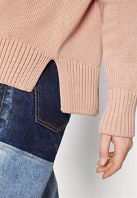 Helles rosa gestricktes Sweatshirt mit geripptem Saum und Bündchen, mit seitlichen Schlitzen. Kombiniert mit dunkelblauen und hellblauen Jeans.
