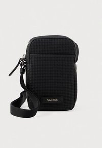 Mustast kootud nailonist risti kantav kott tõmblukuga, reguleeritava rihmaga ja kullakarva "Calvin Klein" logoga plaadiga.