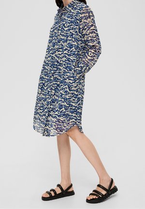 Femme portant une robe chemise à motifs bleu et beige, longueur genou, manches longues, et des sandales noires à brides, debout devant un fond uni.