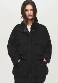 Zwarte oversized denim jacket met twee grote voorkruiszakken, een hoge kraag en knoopdetails. De stof lijkt stevig met een lichte textuur.