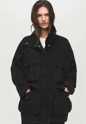 Veste en denim noire oversize avec deux grandes poches avant, un col haut et des détails de boutons. Le tissu semble robuste avec une légère texture.