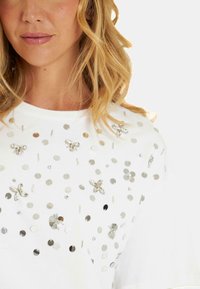 T-shirt en coton blanc avec des sequins métalliques et des embellissements floraux, présentant un motif texturé sur le tissu.