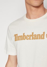Fehér pamut póló, amelyen egy nagy barna "Timberland" logó látható a mellkason. Kerek nyakú és rövid ujjú.