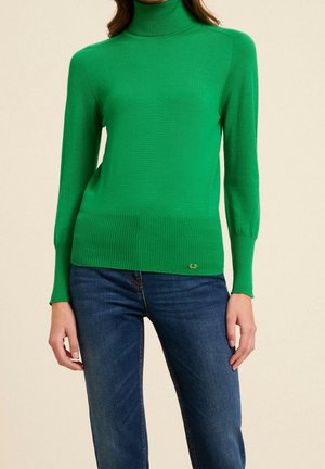 Vrouw met een felgroene coltrui en donkerblauwe spijkerbroek, staand tegen een effen beige achtergrond.