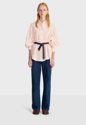 Femme portant une chemise oversize rose clair avec une ceinture tissée, un jean large brodé bleu foncé et des chaussures marron, debout sur un fond blanc.