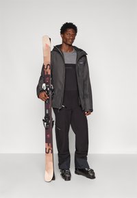 Čierna lyžiarska bunda s šedými akcentmi, šedý dlhý rukáv, čierne lyžiarske nohavice, drží hnedé lyže s viazaním, stojí na neutrálnej pozadí.