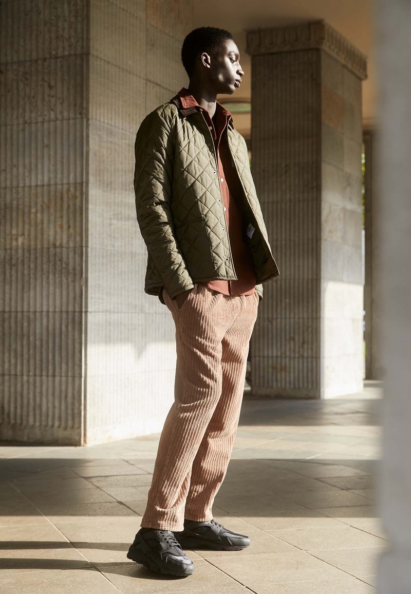 Gepolsterte olivgrüne Jacke über einem rostfarbenen Hemd, kombiniert mit ribbeligen beigen Hosen und schwarzen Sneakers, in einem gut beleuchteten architektonischen Umfeld.