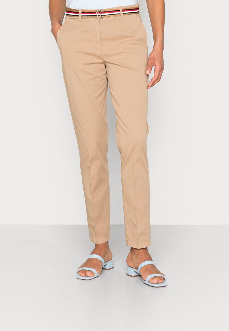 Person som bär beige slim-fit byxor med ett röd, vitt och svart randigt bälte och ljusblå klacksandaler, står med händerna i fickorna.