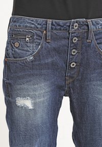 Vaqueros de mezclilla azul oscuro con cintura alta, bragueta de cinco botones, bolsillos laterales y un parche desgastado en el muslo izquierdo.