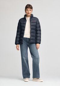 Mörkblå puffjacka med hög krage, quiltat mönster och röd logotyp på bröstet, i kombination med ljusblå vida jeans och beige sneakers.