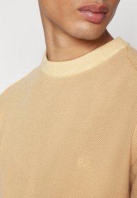 Světle béžový pletený svetr s žebrovaným kulatým výstřihem. Výrazné logo na hrudi v odpovídající barvě, s texturovaným povrchem.