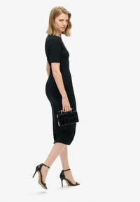 Zwart getailleerd jurkje met korte mouwen en een lengte tot de knieën. Model houdt een kleine zwarte tekstuur clutch vast en draagt zwart gespschoenen.