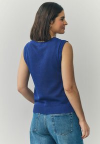 Without sleeves, sinine kudum särk ribaga kaeluse ja äärmise äärega. Kleiditud helesiniste teksadega, esitledes lähedalt liibuvat disaini ja sujuvat tekstuuri.