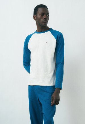 Mand iført hvid og blå langærmet skjorte med matchende blå bukser, stående mod en ensfarvet lys baggrund, kigger sidelæns.