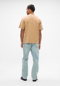 Καφέ βαμβακερό μπλουζάκι, κοντά μανίκια, άνετη εφαρμογή, σε συνδυασμό με ανοιχτό μπλε ίσια τζιν παντελόνια και μαύρα παπούτσια, απεικονιζόμενο από πίσω.