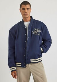 Granatowa kurtka bombowa z pikowaną fakturą, z białym i beżowym paskiem na dolnym brzegu i mankietach oraz wyszywanym akcentem z literą "M".