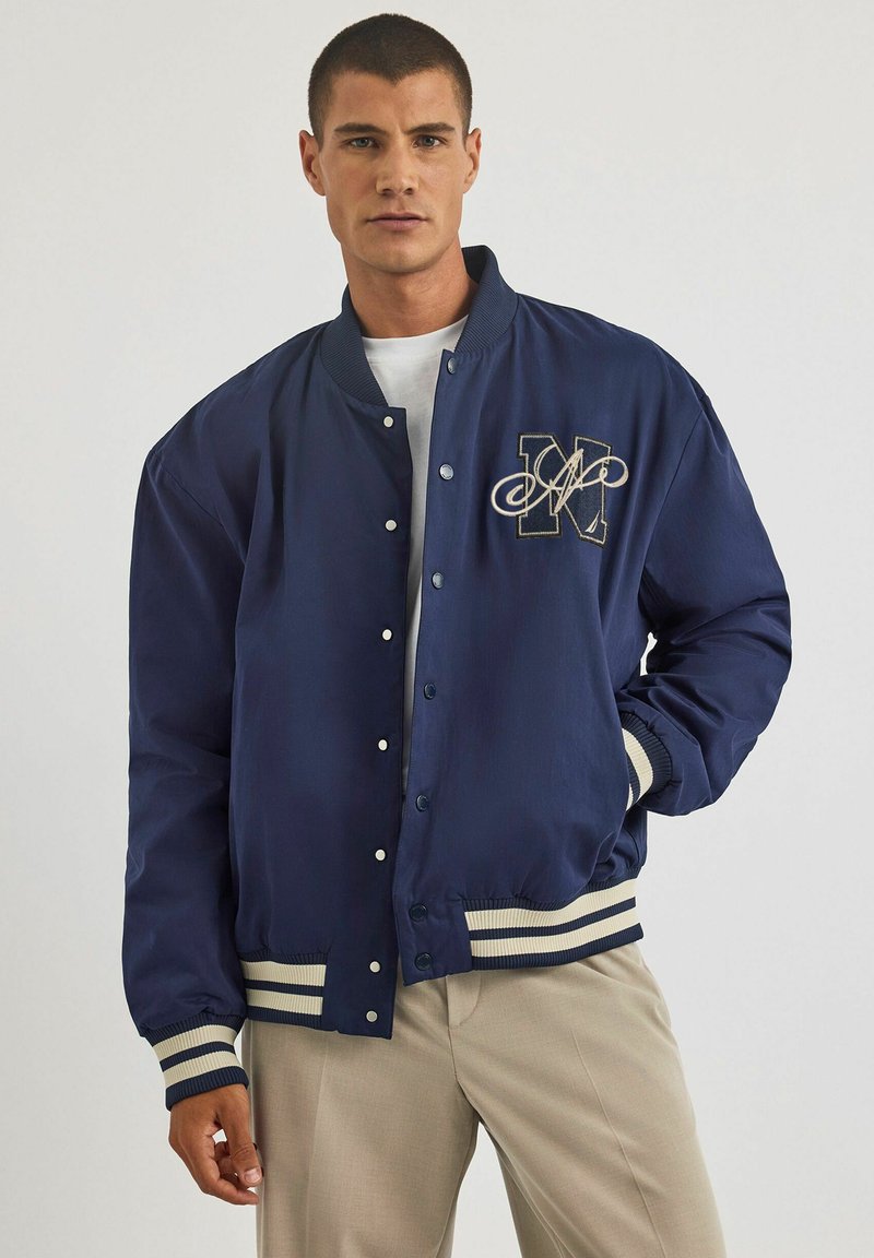 Granatowa kurtka bombowa z pikowaną fakturą, z białym i beżowym paskiem na dolnym brzegu i mankietach oraz wyszywanym akcentem z literą "M".