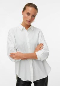 Chemise blanche à boutons avec col, manches longues et coupe légèrement ample. Présente des accents de perles discrets et une texture lisse.