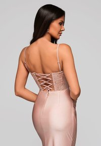 Femme aux longs cheveux foncés portant une robe en satin rose dos nu avec un corset lacé et des bretelles fines, tournée de profil sur un fond uni.