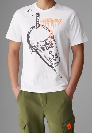 Hombre con camiseta blanca con un diseño en estilo aerosol negro y naranja de un vaso de bebida volcado y texto "FIRE + ICE" y pantalones cargo de color verde oliva.
