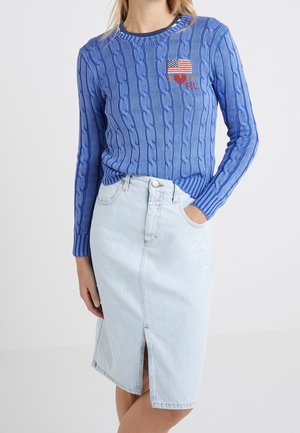 Femme portant un pull en maille torsadée bleu avec un patch drapeau américain et cœur, associé à une jupe en jean bleu clair avec une fente à l'avant.