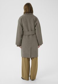 Abrigo de lana gris con cintura ceñida, mangas amplias y textura suave. La modelo lo combina con pantalones beige de pierna ancha.