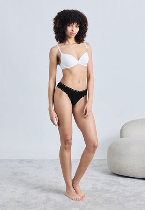 Femme aux cheveux bouclés, debout pieds nus sur un tapis, portant un soutien-gorge blanc et une culotte noire, avec une chaise moderne gris clair en arrière-plan.