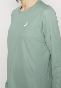 Langärmliges Sportshirt aus leicht grünem Stoff mit glatter Textur, das auf der Brust ein kleines weißes Logo und minimale Nähte aufweist.