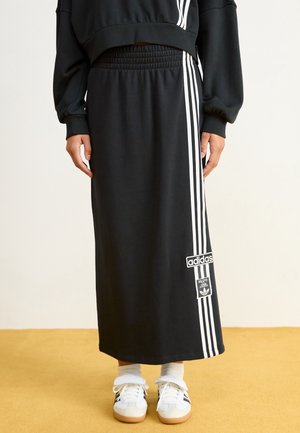 Sort Adidas nederdel med hvide tre striber ned langs siden, elastisk taljebånd, kombineret med hvide sneakers og sokker på beige gulv.