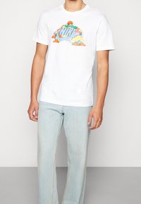 Camiseta blanca de algodón con un diseño gráfico colorido que incluye nubes, árboles y una cara sonriente, combinada con unos jeans de denim azul claro.