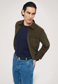 Oliwkowa koszula zapinana na guziki na granatowej koszulce, połączona z jasnoniebieskimi dżinsami i ciemnozielonym paskiem z srebrną klamrą.