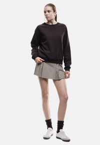 Sudadera marrón oscuro con mangas raglán, combinada con una falda plisada de cuadros beige. La modelo lleva zapatillas blancas y calcetines oscuros.