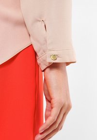 Blusa beige chiaro con maniche arrotolate, caratterizzata da un accento di bottone dorato. La blusa è abbinata a un indumento rosso brillante.