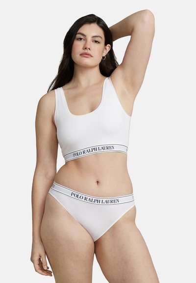 Femme portant un bralette Polo Ralph Lauren blanc et des sous-vêtements assortis, posant avec un bras levé sur un fond clair uni.
