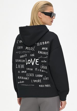 Persoon die een zwarte hoodie draagt met "love" in meerdere talen op de achterkant geschreven, gecombineerd met een grijze broek en zwarte zonnebril.
